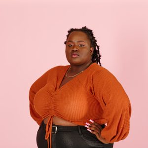 partner 1 Young confident black plus size body positive woman looks at camera holding hands on waist on light pink background in studio closeup